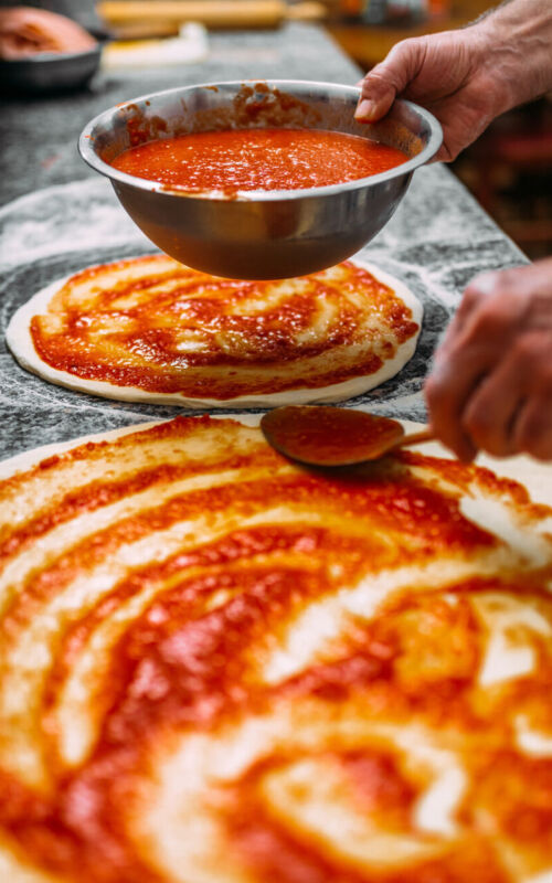 Chef baker making pizza at the kitchen. Adding tomato sauce on a pizza dough.